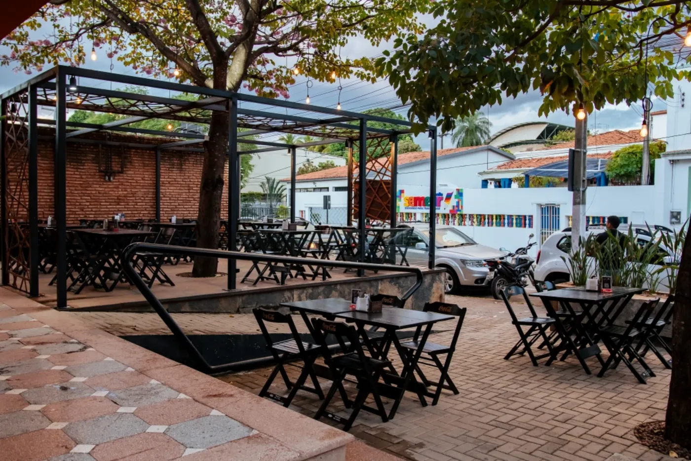 Área externa de restaurante com pergolado metálico, varal de lâmpadas e muro de tijolo aparente